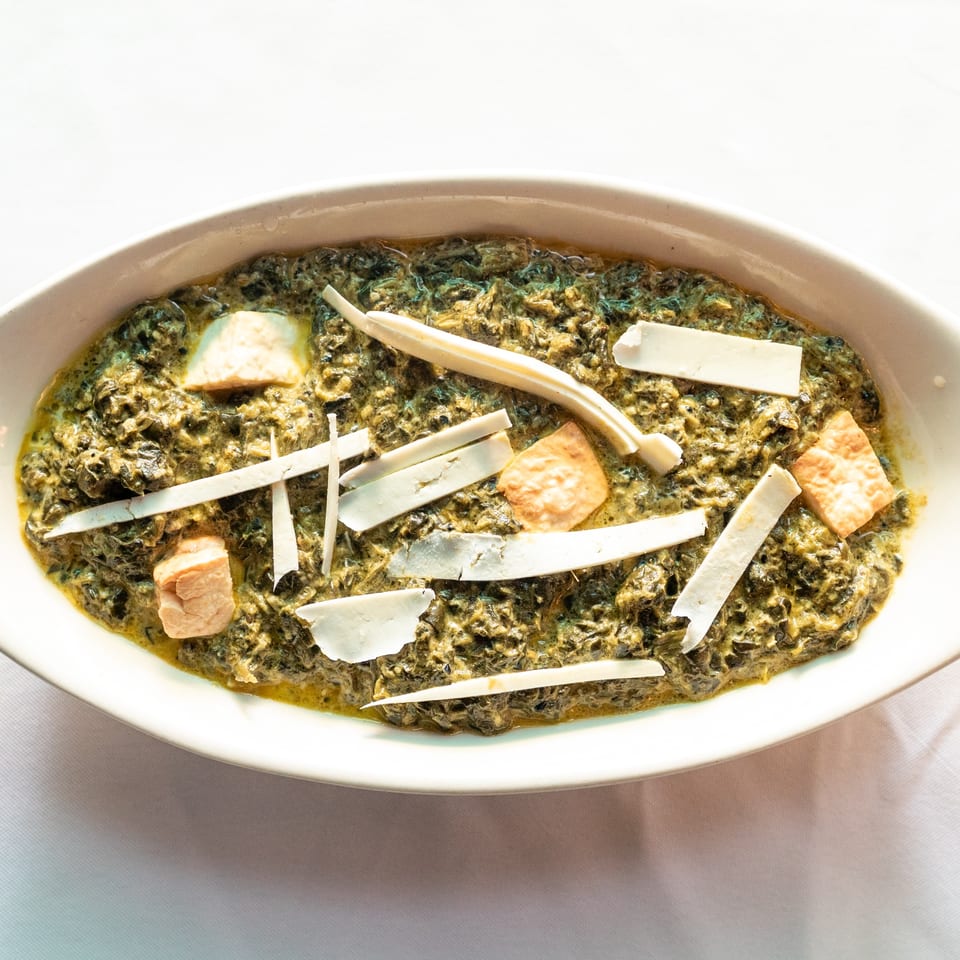 A white oval bowl filled with green saag paneer, garnished with cheese slices and cubes.
